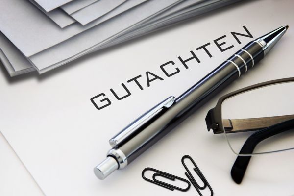 Das Wort Gutachten auf weißem Papier, darauf liegt ein Kugelschreiber, Büroklammern und eine Brille